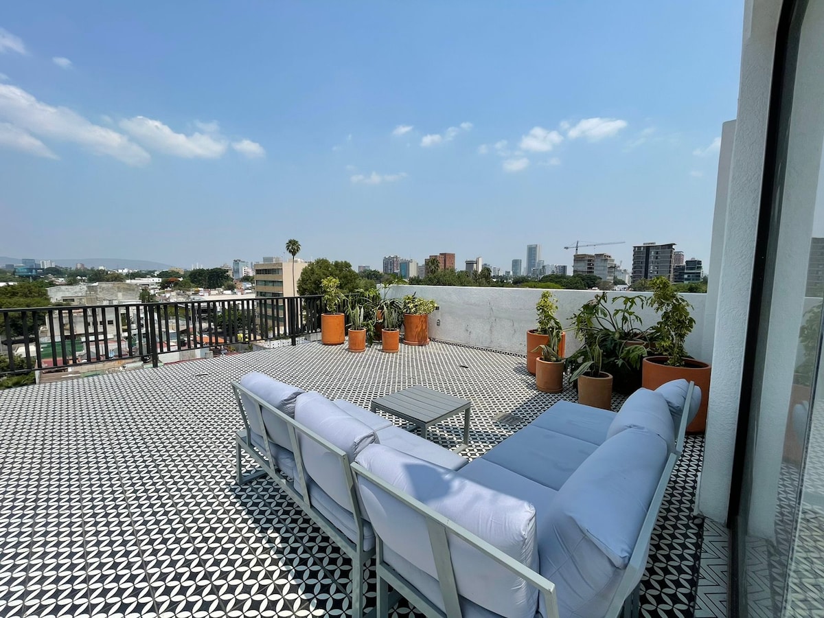 A spacious rooftop terrace is presented, featuring a comfortable white sofa and a small table. Potted plants are arranged around the area, providing greenery against a backdrop of urban architecture and a clear blue sky.