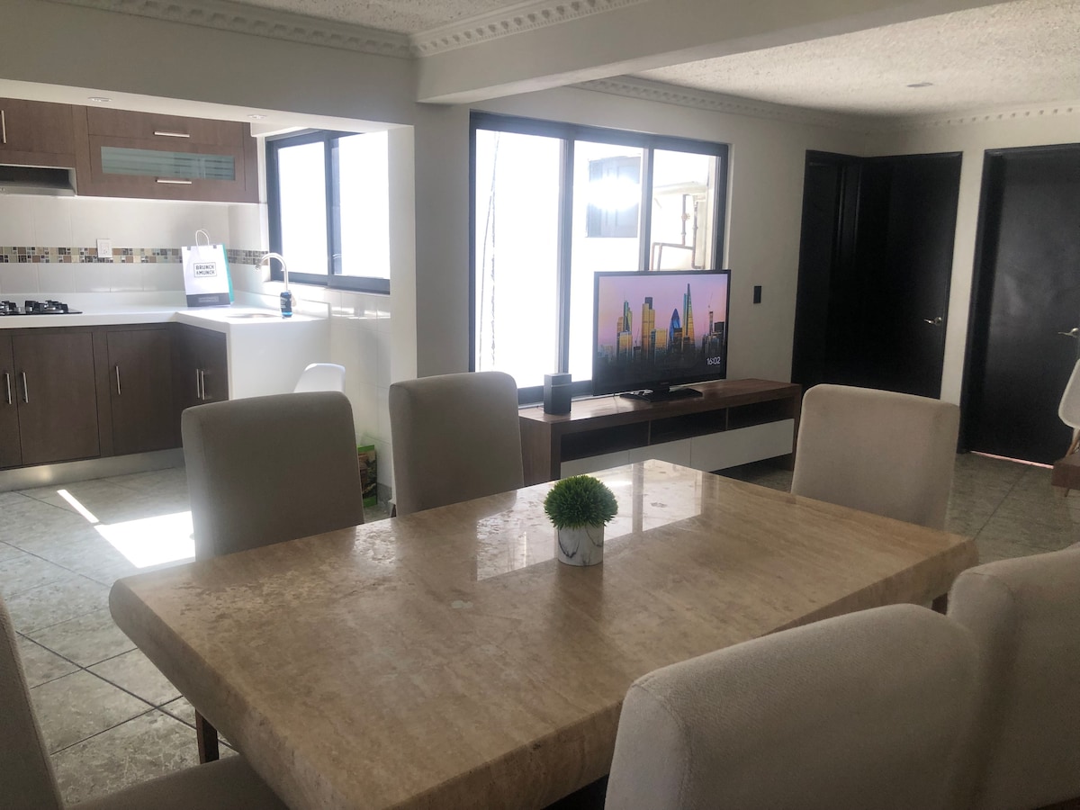 A modern shared living space features a large dining table surrounded by upholstered chairs. A flat-screen television is positioned on a media console. Large windows provide natural light, illuminating the open kitchen area with contemporary cabinetry and appliances.
