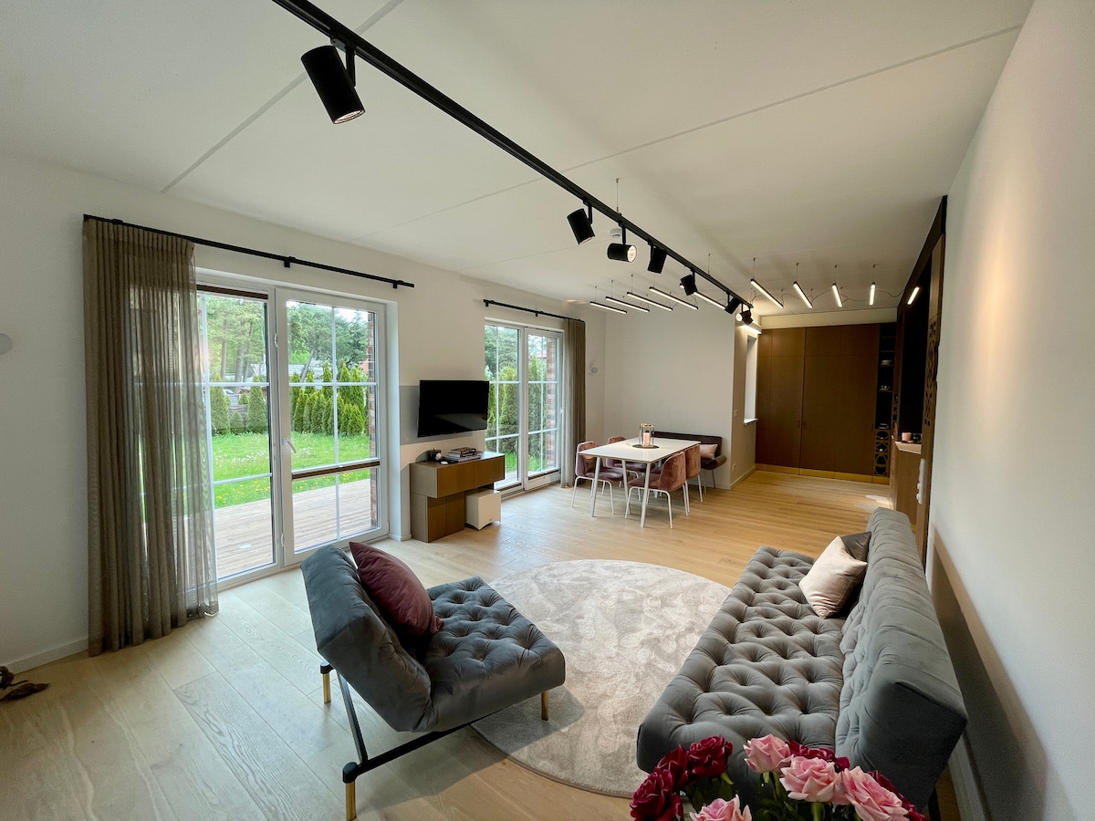 The spacious living area features a light color palette with large glass doors opening to a green outdoor space. Stylish seating includes two comfortable sofas and a round rug, while a dining table is set for meals. Soft natural light enhances the inviting atmosphere.