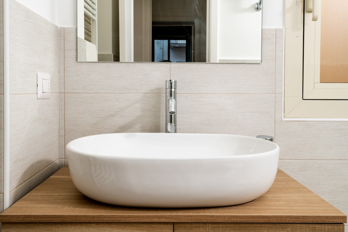 A modern bathroom sink is displayed on a wooden countertop. The sleek, oval-shaped basin is paired with a minimalist faucet. A mirror is mounted above the sink, reflecting the neutral tones of the walls and nearby window.