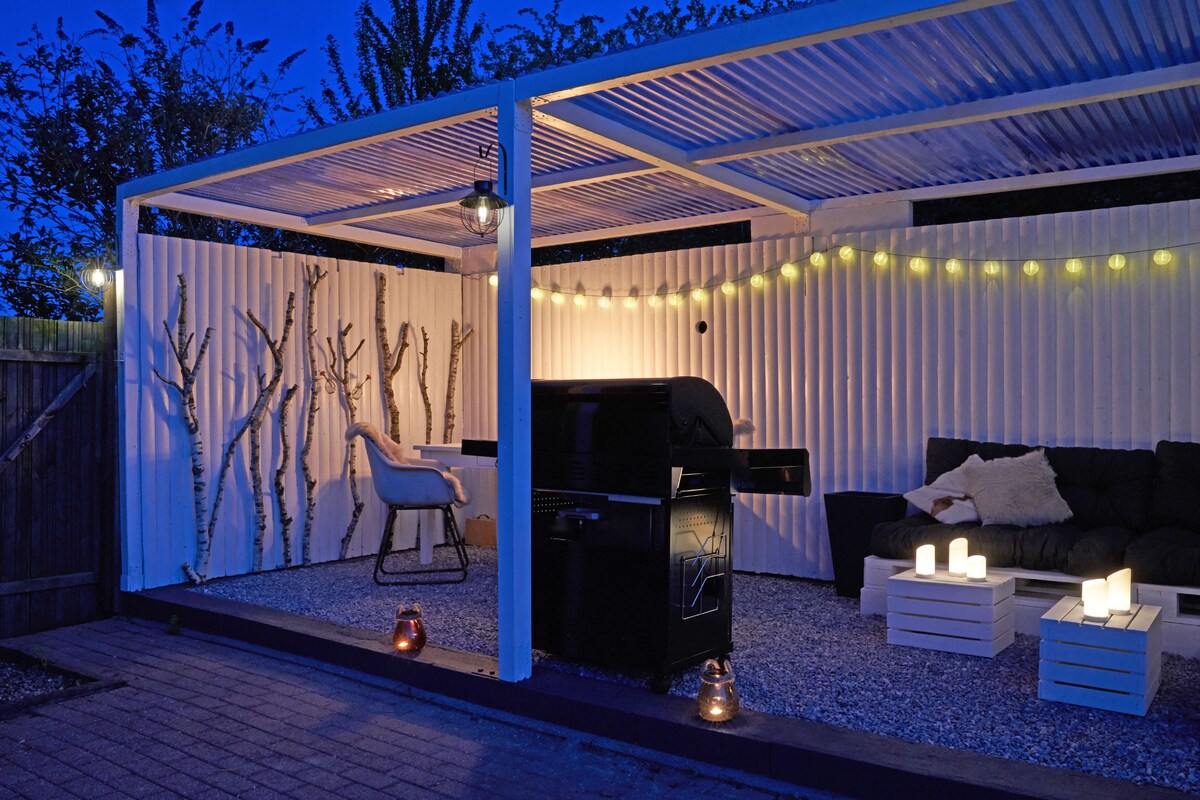 An outdoor space is presented with a covered area featuring a black grill, cozy seating, and soft lighting. The gravel floor is complemented by white lanterns and a wood-textured wall with tree branches. String lights enhance the inviting ambiance for evening gatherings.