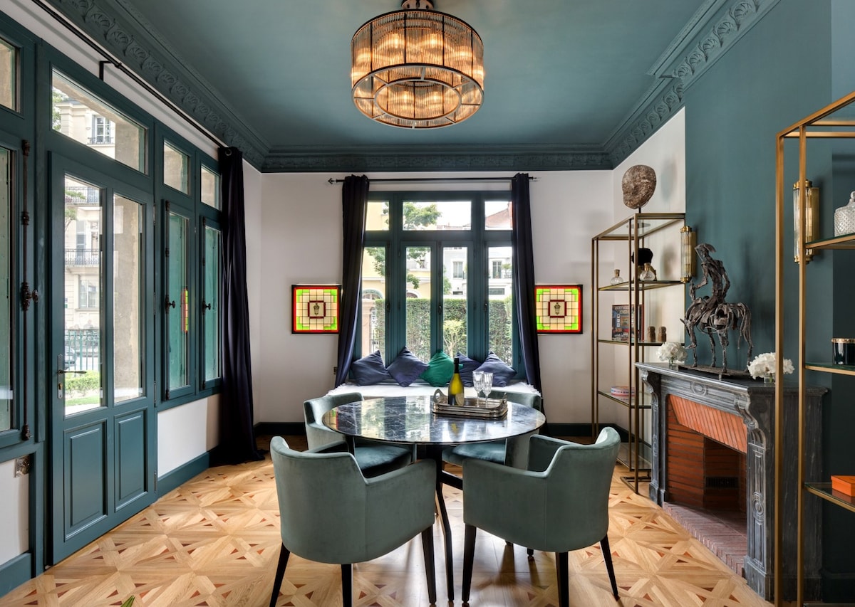 A stylish dining area is showcased with a round marble table surrounded by four plush green chairs. Large windows framed in blue allow natural light to fill the space, highlighting the intricate flooring and elegant lighting fixture above.