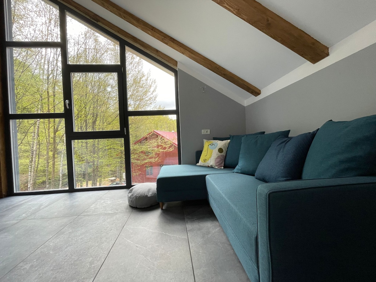 A spacious living area features a comfortable sectional sofa in teal, complemented by a decorative cushion. Large windows allow natural light to fill the room and frame views of the surrounding greenery, enhancing the connection with the outdoors.