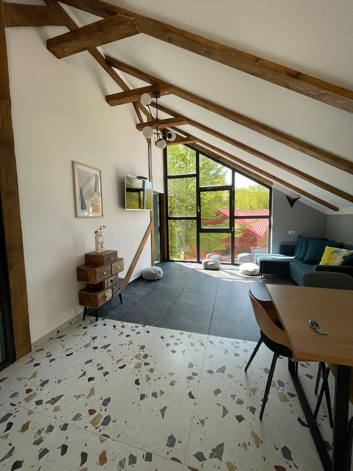 An open living area features sloped wooden ceilings and large glass windows offering natural light. A stylish green sofa is positioned near a sleek wooden sideboard, and a dining table is set for meals. The floor showcases unique speckled patterns, contributing to the room's character.