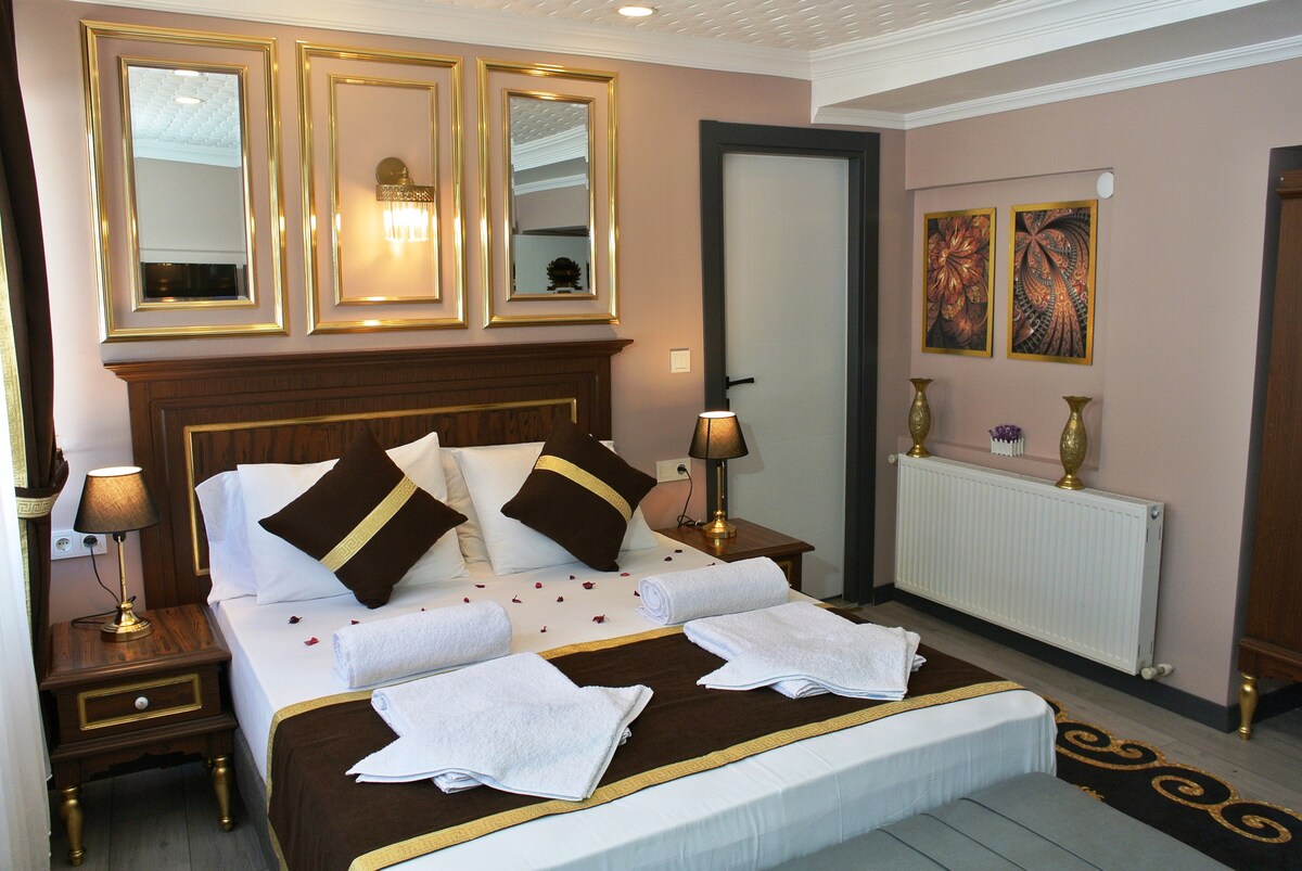 A well-appointed room features a queen-size bed adorned with decorative pillows and two rolled towels. Stylish lamps are positioned on either side of the bed. Decorative artwork and mirrors enhance the room's elegance. A radiator and door are visible in the background.