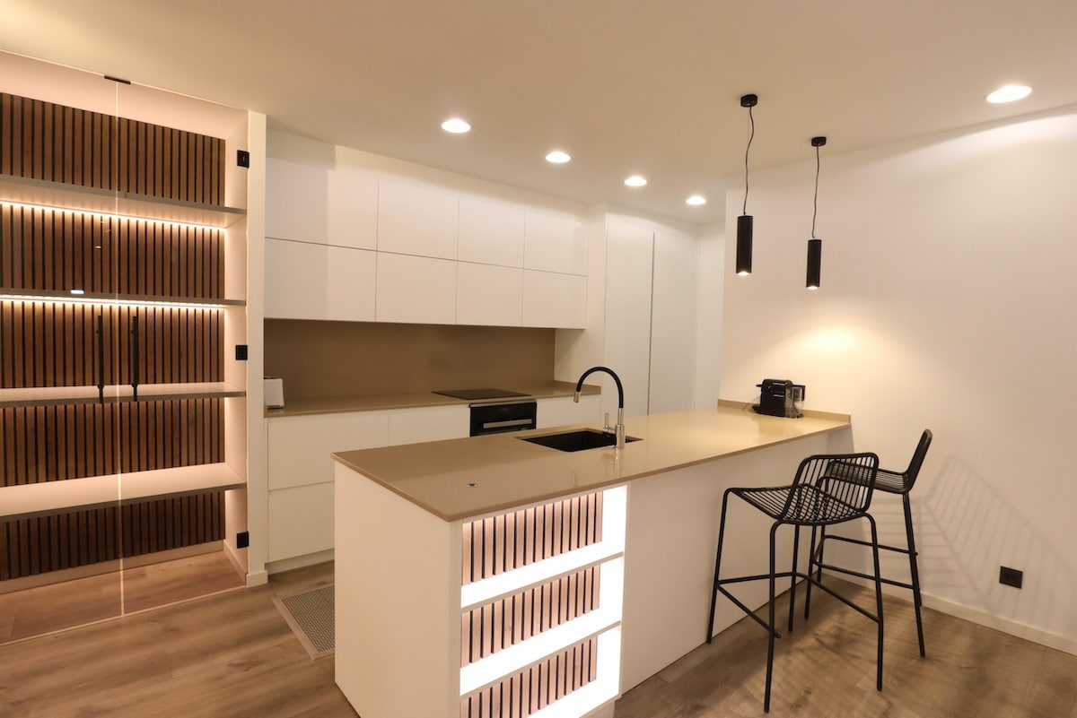 A modern kitchen features sleek white cabinetry and a large central island with a contrasting light countertop. Subtle lighting highlights the textured wooden wall paneling, while black pendant lights add a contemporary touch. A minimalist bar stool is positioned at the island for casual seating.