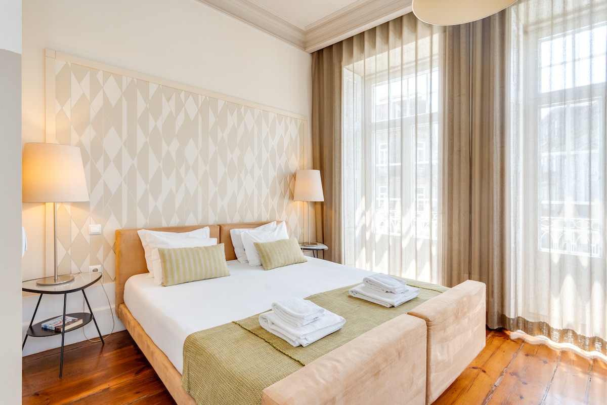 A spacious bedroom features a queen-size bed with a light-colored headboard, complemented by decorative pillows. Two lamps provide soft lighting, and large windows with sheer curtains allow natural light to fill the room. Fresh towels are neatly arranged at the foot of the bed.