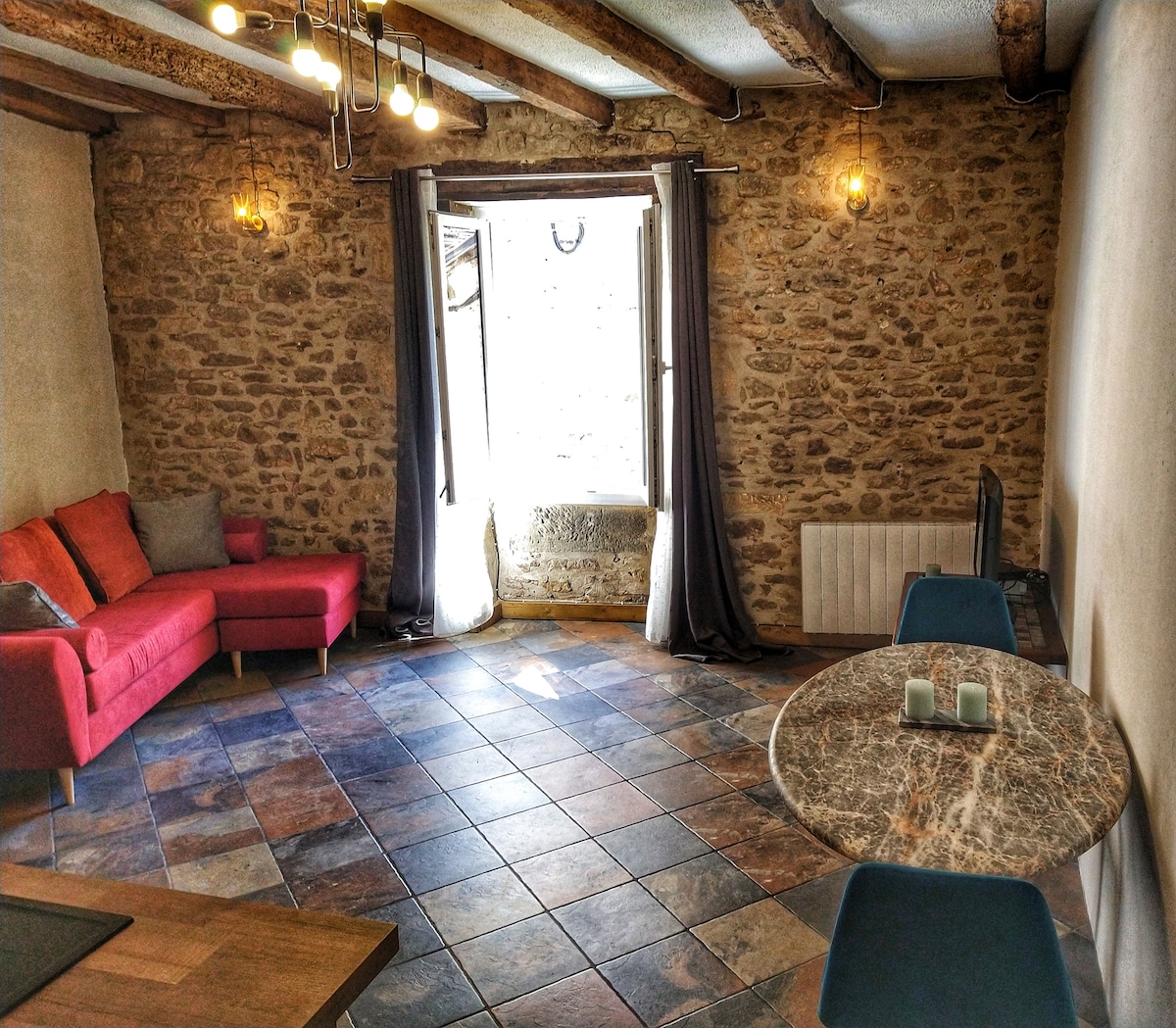 A spacious living area is highlighted by exposed stone walls and wooden beams. A red sofa adds a pop of color, while a small round table with blue chairs sits nearby. Natural light enters through a window, enhancing the inviting environment.
