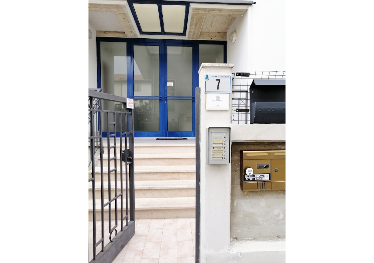 The entrance features a set of stone steps leading up to a glass door framed by blue accents. A gate is visible to the left, along with a mailbox and intercom mounted on the wall beside the door.