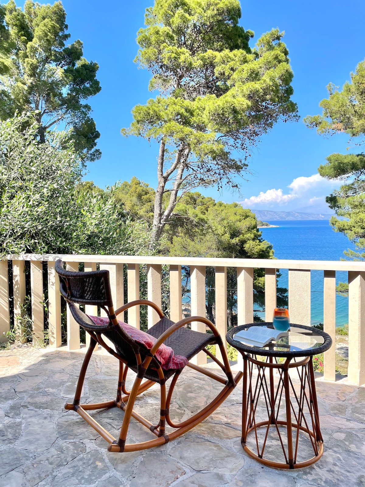 A rocking chair is positioned on a stone terrace, offering serene views of the sea and surrounding lush greenery. A small, round table stands nearby, complementing the relaxing outdoor space, which is surrounded by tall trees and features a clear blue sky in the background.