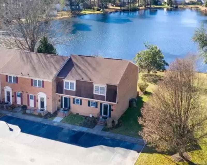 An aerial view captures a two-story condo set among tidy landscaping. The backdrop features a serene lake surrounded by trees, reflecting the sky. The neighboring units display a mix of brick and siding, with a private patio visible on the side.