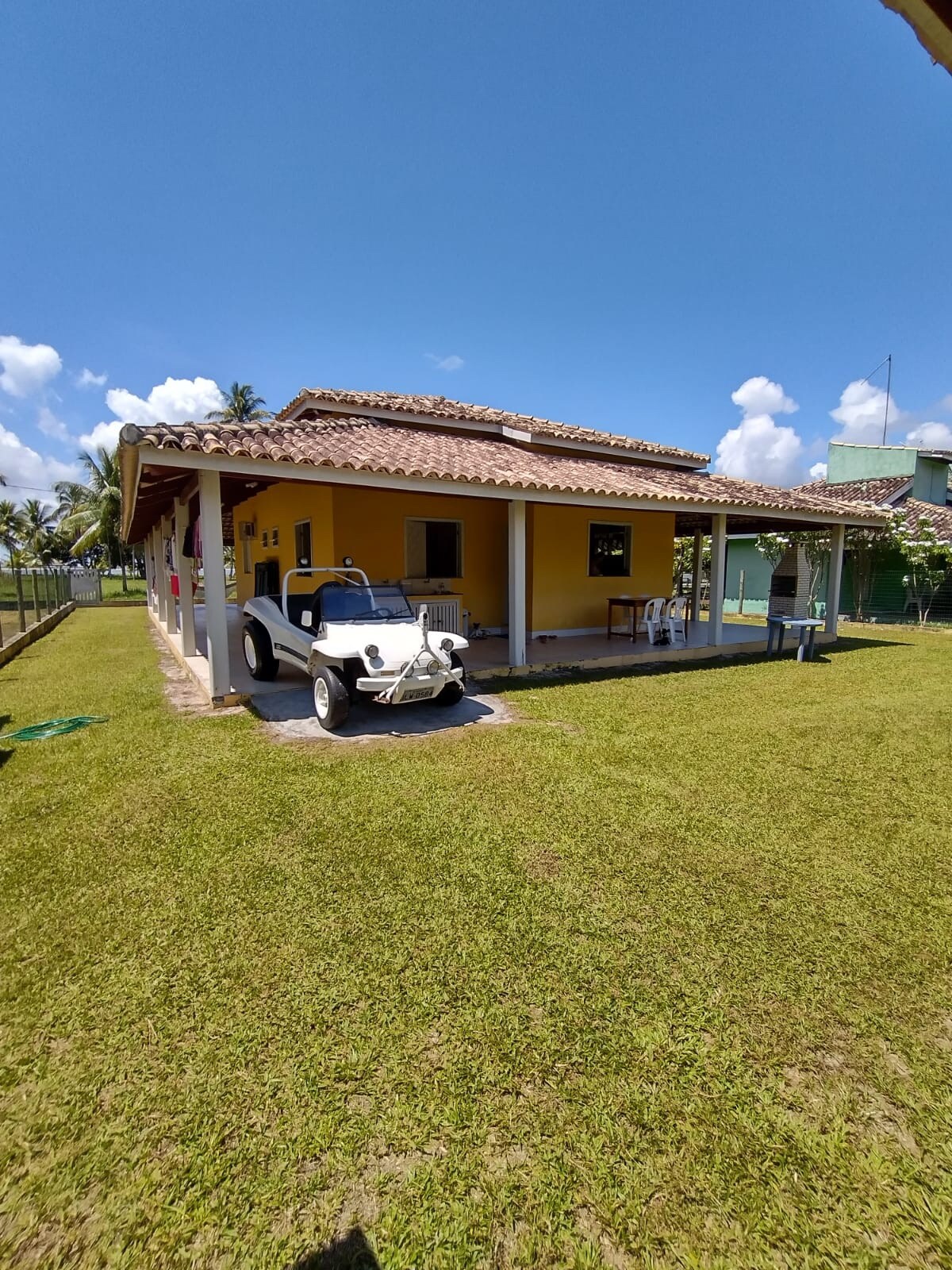 The exterior of a vibrant yellow house is highlighted under a clear blue sky. A covered porch is visible, accompanied by a classic white vehicle parked nearby. Lush green grass surrounds the property, with palm trees and other greenery in the background.