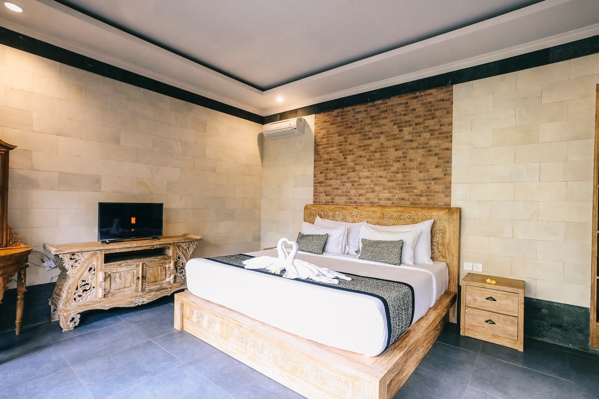 A spacious bedroom features a large wooden bed with neutral bedding and decorative pillows. An intricately carved wooden cabinet with a television sits across from the bed. Natural stone walls enhance the calming atmosphere, while air-conditioning units are discreetly positioned to maintain comfort.