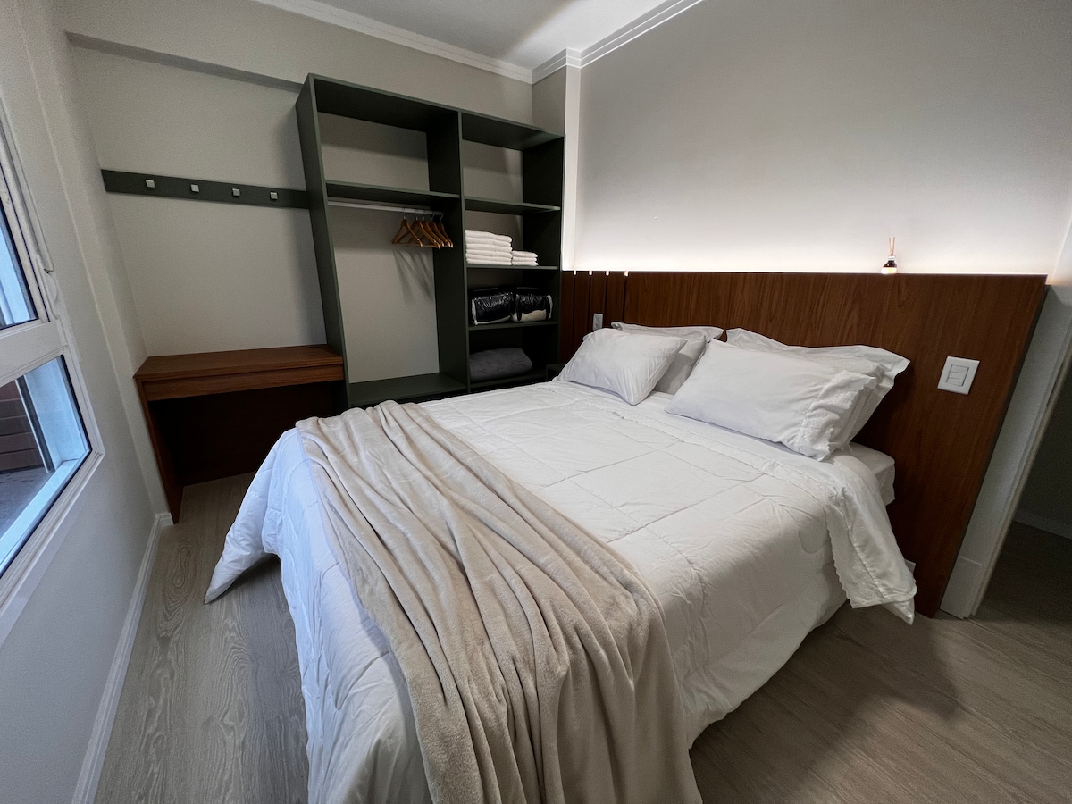 A serene bedroom features a spacious bed with white linens and a light throw draped over it. A wooden headboard and accompanying nightstands create a calming aesthetic. Storage shelving is visible alongside a window, providing natural light and a cozy atmosphere.