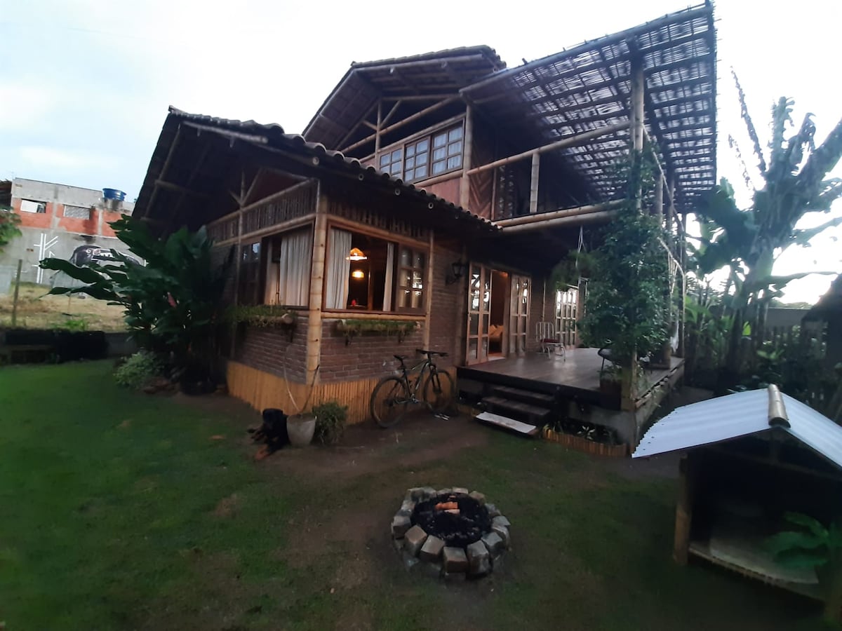 A spacious bamboo house is set amidst lush greenery, featuring a large porch with ample seating. The exterior showcases natural materials, with tall windows and an overhanging roof. A fire pit is visible in the yard, surrounded by garden space. Bicycles are parked near the entrance.