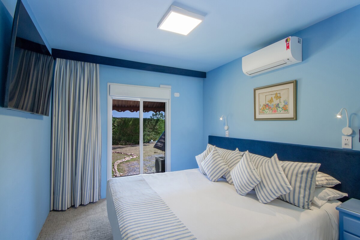 A bright room is designed with calming blue walls, featuring a spacious bed adorned with striped pillows. A flat-screen TV is mounted on the wall, while light curtains frame a sliding door that opens to a scenic outdoor area.