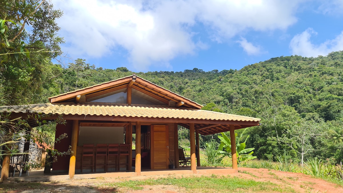 The private chalet features a wooden structure with a sloped, tiled roof. Large windows provide views of the surrounding lush greenery. A spacious porch area is visible, adorned with wooden furniture, inviting relaxation in nature.