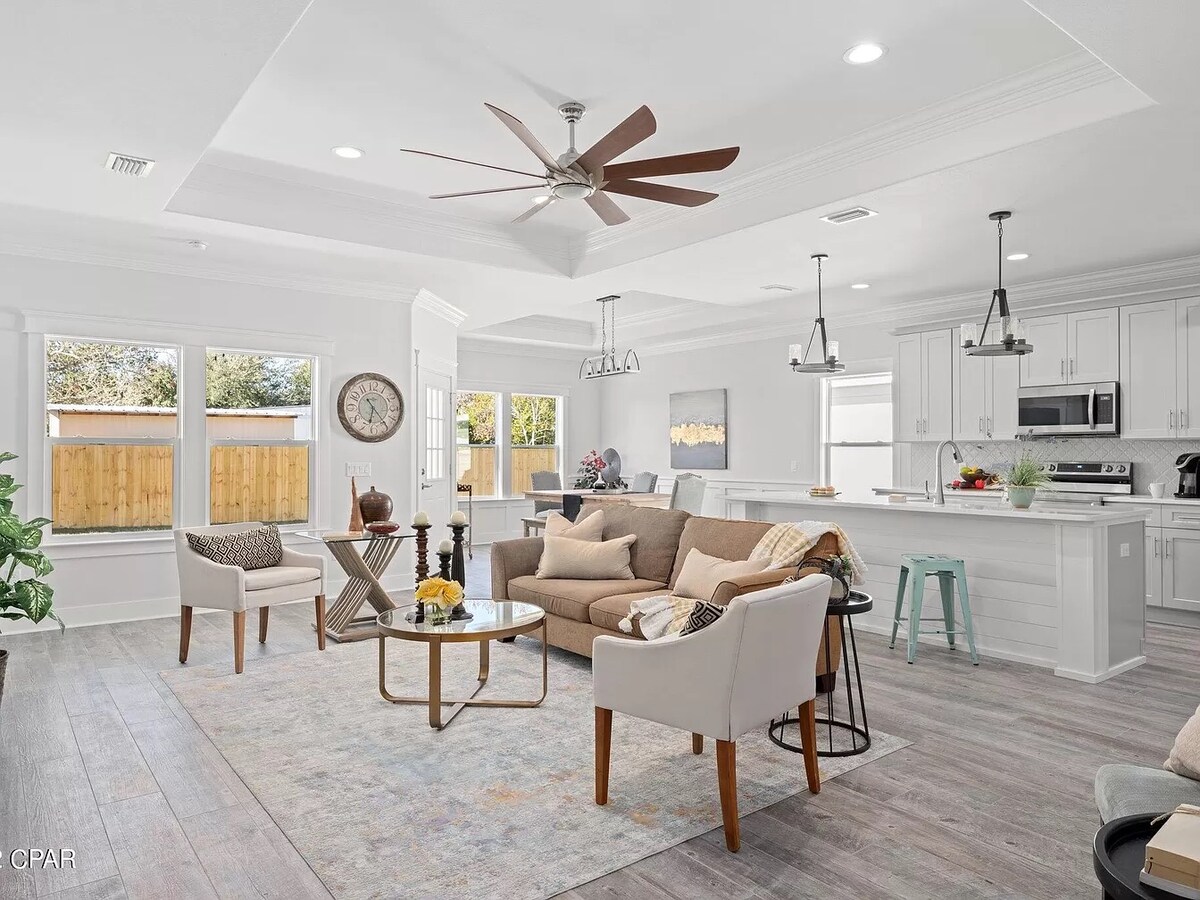 The open living area features a modern design with high ceilings and ample natural light. A comfortable seating arrangement includes a lounge sofa and two armchairs, centered around a coffee table. A fan hangs above, and the kitchen is visible, showcasing white cabinetry and bar seating.