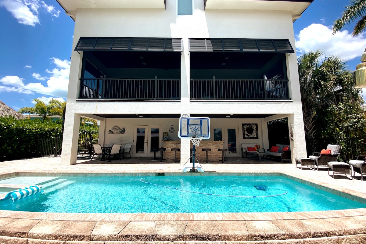 The outdoor area features a heated pool, with a basketball hoop positioned nearby. Sun loungers are arranged around the pool, and the space is shaded by the elevated patio above, which includes several seating options for relaxation.