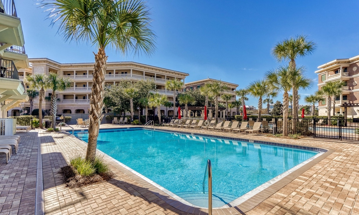 A spacious outdoor pool area is surrounded by tall palm trees and lounge chairs. The water is clear and inviting under a bright blue sky, with a building visible in the background that showcases a tropical aesthetic.