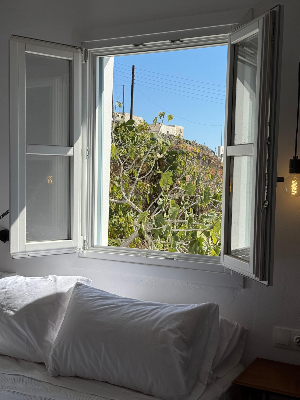 Large white-framed windows are opened to reveal a vibrant view of greenery outside. Sunlight streams into the airy space, illuminating the cozy sleeping area adorned with soft white bedding.
