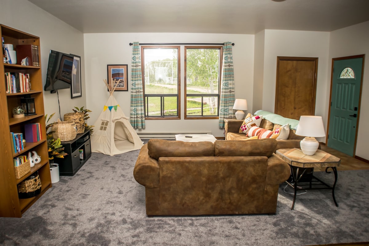 A cozy living room features a brown sectional sofa, a woven basket, and a coffee table. Large windows provide a view of the outdoor landscape. A bookshelf is filled with books, and a play tent adds a playful touch to the inviting space.