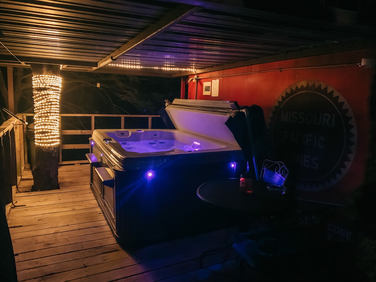 An outdoor hot tub is illuminated by blue lighting, positioned on a wooden deck beneath a canopy. Soft string lights enhance the ambiance, with a small table and chair set nearby. The backdrop features a vintage Missouri Pacific logo displayed on the wall.