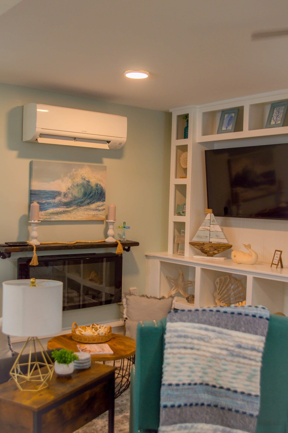 A living room space features a mounted flat-screen television above a decorative fireplace. A painting of ocean waves is displayed on the wall. Bookshelves filled with seashells and beach-themed decor surround the television. A cozy seating area includes a patterned throw and a stylish lamp.