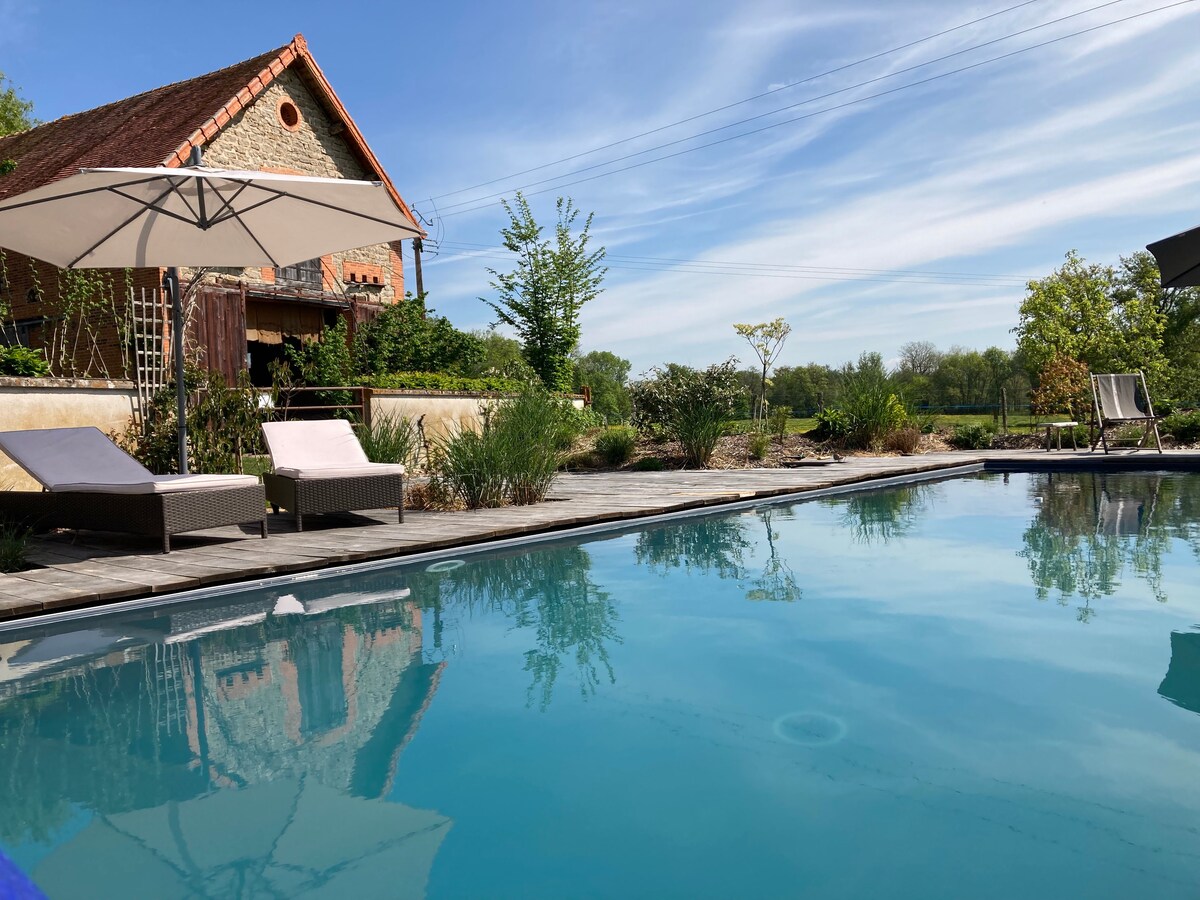 A serene outdoor pool area is highlighted, featuring sleek lounge chairs beside the clear water. Umbrellas provide shade, while lush greenery and garden plants surround the pool, enhancing the tranquil environment. The charming home is visible in the background under a bright blue sky.