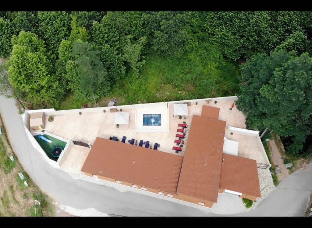 An aerial view captures a single-story house surrounded by lush greenery. A spacious patio features a jacuzzi-pool, lounge chairs, and shaded areas. The layout showcases a harmonious blend of outdoor and indoor spaces, with a clear view of the surrounding natural environment.