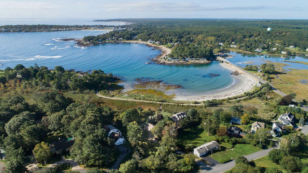 An aerial view showcases a serene coastal landscape, featuring sandy beaches along the shoreline, lush greenery surrounding the bay, and a winding road leading into the residential area. Clear blue waters reflect the sky under a calm atmosphere, accentuated by gentle waves along the shore.