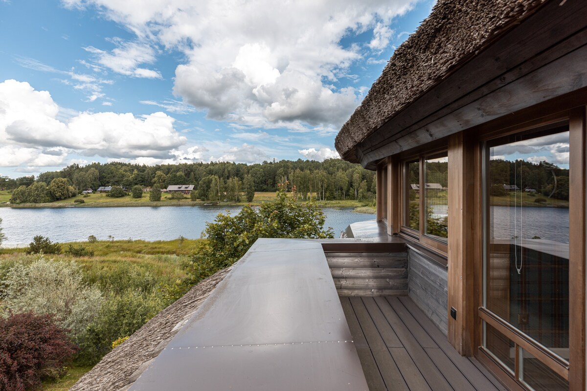 A balcony with wooden railings overlooks the serene lake, framed by lush greenery. The sky is dotted with white clouds, reflecting a tranquil atmosphere. The rustic architecture of the building features natural materials, blending harmoniously with the surrounding landscape.
