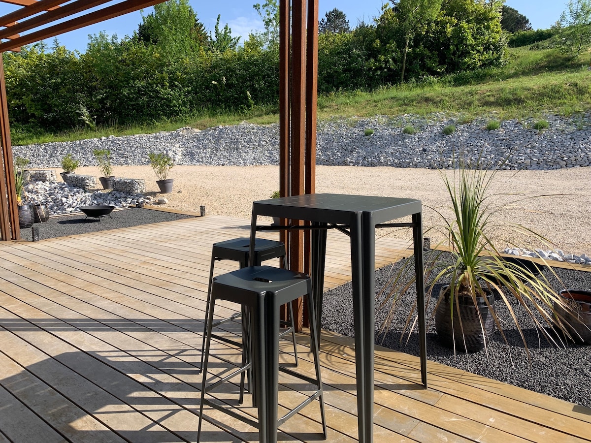 A shaded outdoor terrace features a sleek black table accompanied by two matching stools. Surrounding greenery and gravel landscaping create a serene atmosphere, while potted plants add subtle nature elements to the space.