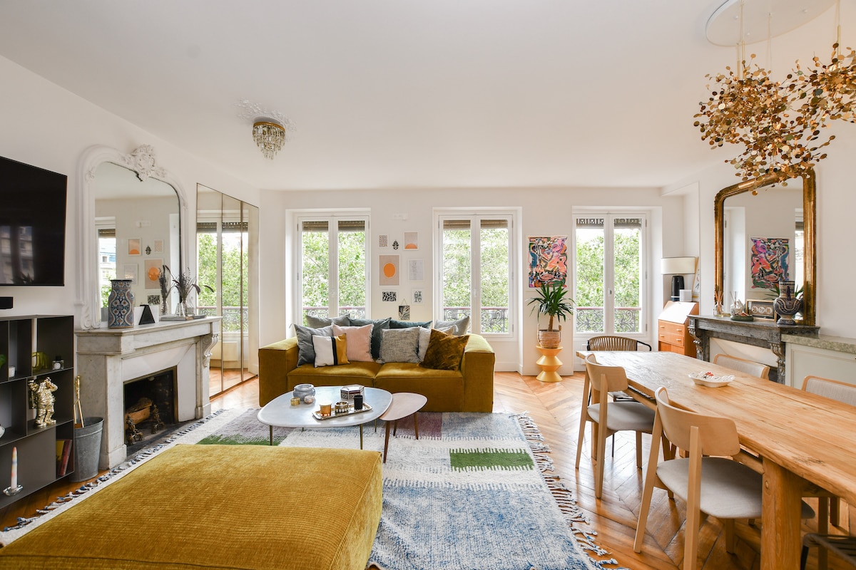 A spacious living area is highlighted by large windows that invite natural light and offer views of greenery. A comfortable mustard-yellow sofa is placed around a low coffee table, with a patterned area rug beneath. A wooden dining table accommodates several chairs, enhancing the convivial atmosphere.