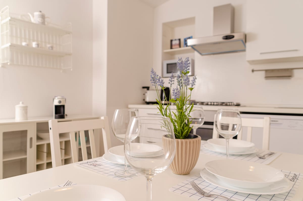 A bright kitchen area is shown, featuring a dining table set with white plates and glassware. A small potted plant with purple flowers adds a touch of color. In the background, modern kitchen appliances are neatly arranged, contributing to a functional space.