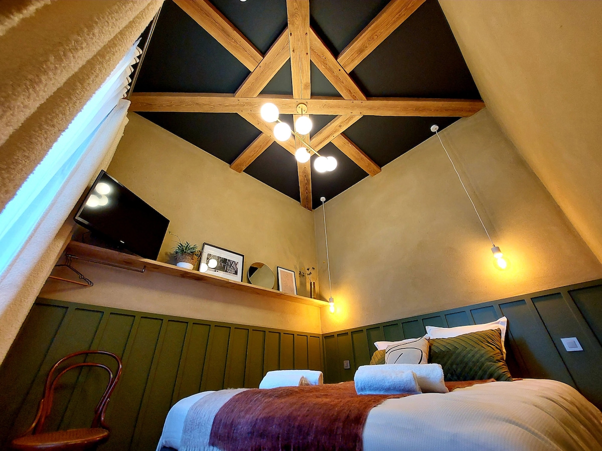 The room features a cozy layout with a spacious bed, accented by natural wood beams overhead. Soft lighting is provided by pendant fixtures, and a television is mounted on the wall. A decorative shelf displays art and plants, enhancing the inviting space.