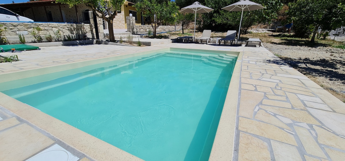A private pool is surrounded by sun loungers and umbrellas on a tiled patio. Lush greenery and a stone wall create a serene outdoor setting. Clear blue water reflects the sky, offering a refreshing escape. Sunlight illuminates the area, enhancing the tranquil atmosphere.