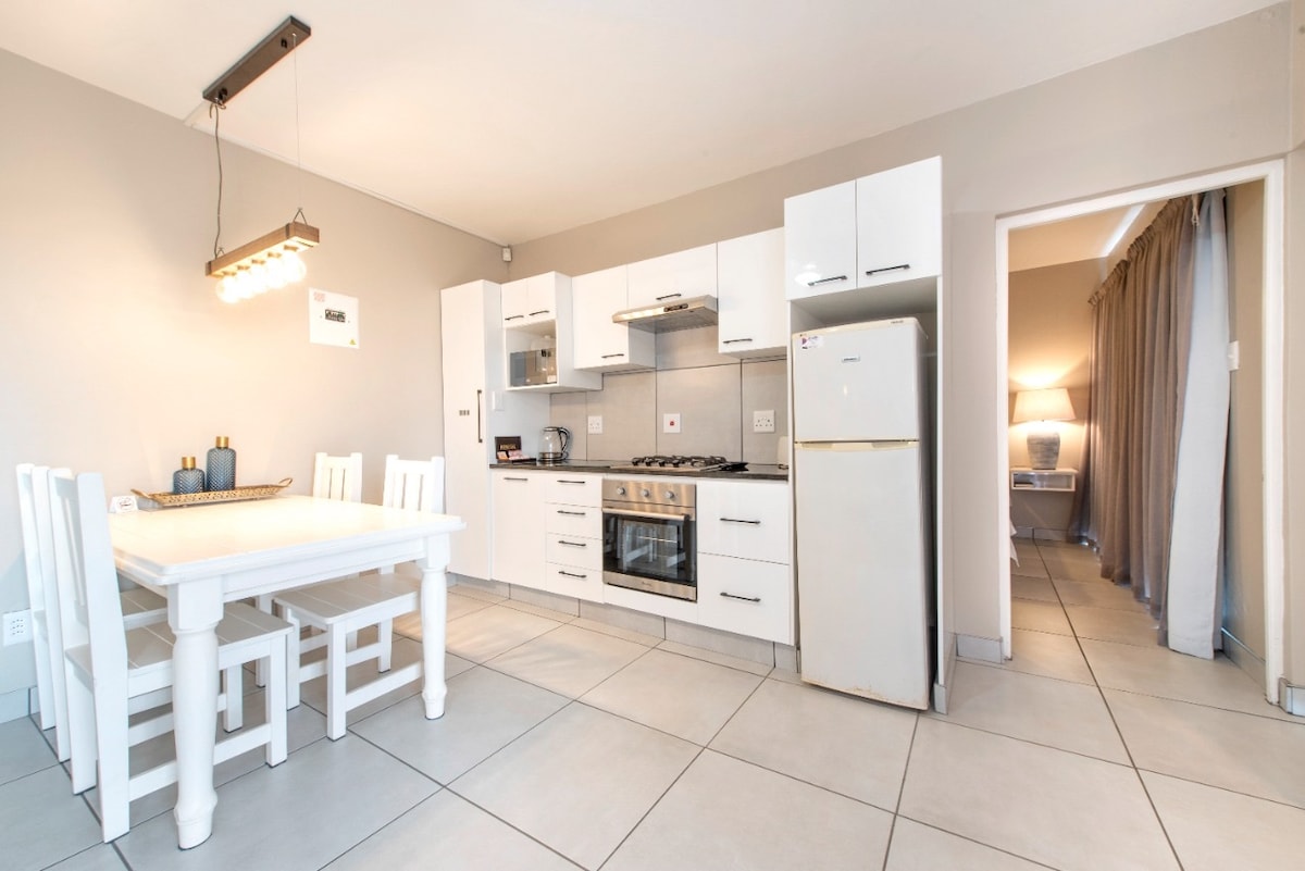 A functional kitchen and dining area features a white wooden table accompanied by four chairs. Cabinets are arranged above and below the countertop, which includes a gas stove and oven. A refrigerator stands nearby, and warm light is provided by a modern overhead fixture.
