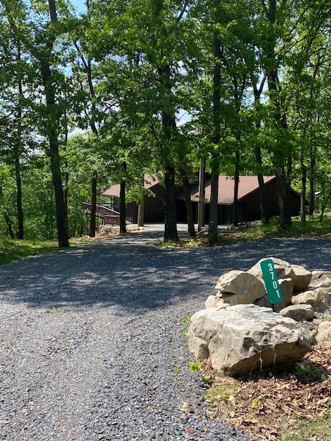 A cabin is visible through a wooded area, set back from a gravel driveway. Towering trees surround the property, providing ample shade. A large rock with the number '3701' is positioned at the edge of the driveway, marking the entrance to this secluded home.