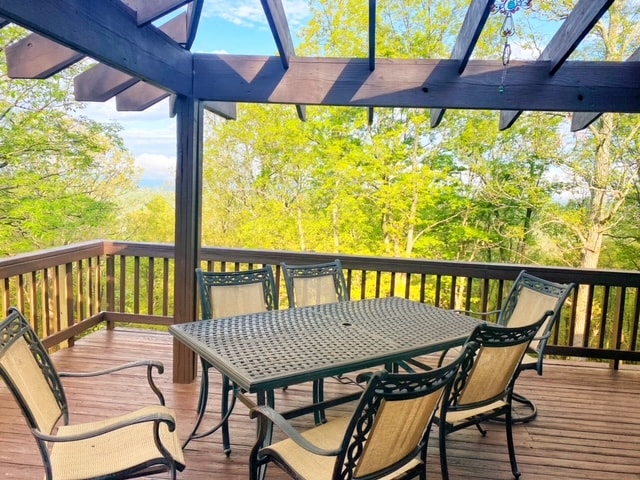 A spacious outdoor dining area is situated on a wrap-around deck, featuring a large metal table surrounded by six cushioned chairs. Overhead, a wooden pergola provides partial shade, with lush greenery visible in the background, enhancing the serene outdoor setting.