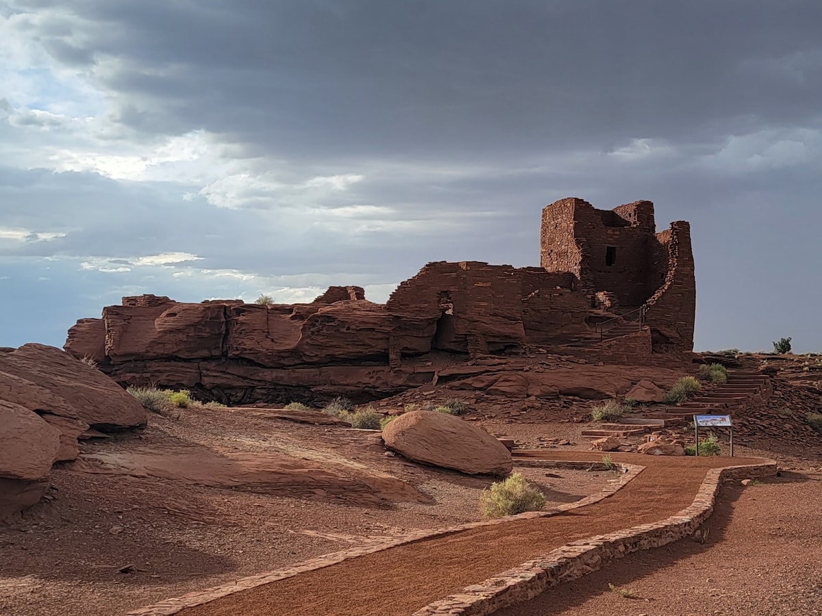 An ancient stone structure sits atop a rocky landscape, surrounded by reddish-brown earth and sparse greenery. A winding path leads toward the ruins, with a cloudy sky providing a dramatic backdrop. The remnants of walls and towers are visible against the natural terrain.