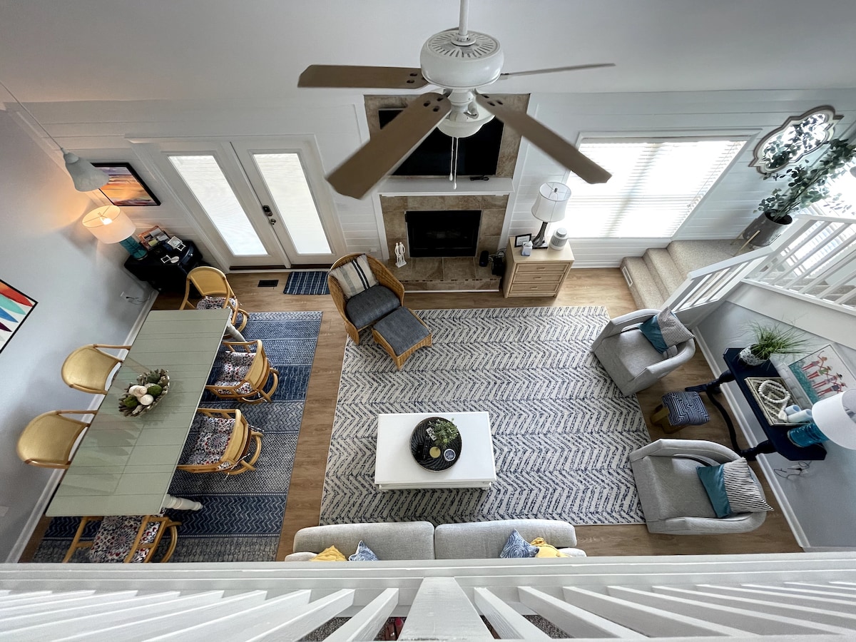 An expansive open floor plan is visible, featuring a dining area with four chairs, a cozy living space with a sectional sofa, and a coffee table. Natural light floods the room through large windows, while a ceiling fan adds comfort. Subtle decor and textures enhance the welcoming atmosphere.