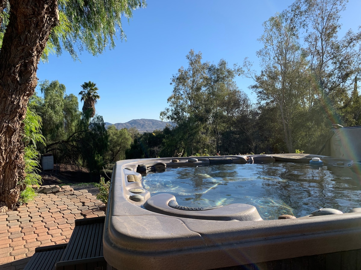 A hot tub is situated on a wooden deck, surrounded by greenery and trees. The view beyond includes rolling hills under a clear blue sky, enhancing the serene outdoor atmosphere.