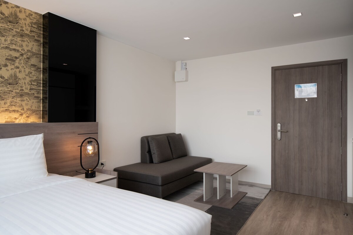 A modern room features a spacious bed with white linens and a wooden headboard. A comfortable sofa is positioned beside a small table. The entry door includes a notice, and muted decor enhances the inviting ambiance.