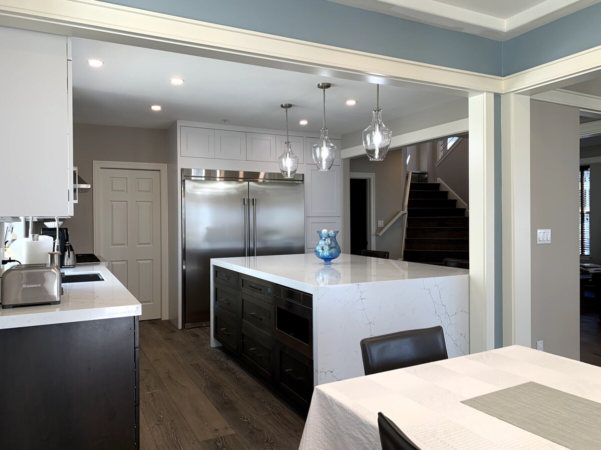 A modern kitchen features a large island with a marble counter, illuminated by pendant lights. Stainless steel appliances are visible, and ample cabinetry provides storage. Light wood flooring complements the neutral color palette, creating a bright and open cooking space.