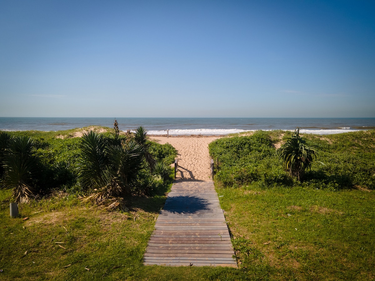 A wooden walkway leads through lush greenery toward a sandy beach. The ocean can be seen in the distance, with gentle waves lapping at the shore under a clear blue sky.