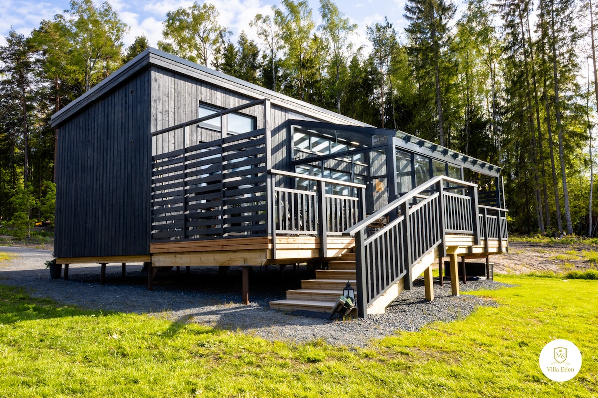 The exterior of Villa Eden is depicted, showcasing a modern wooden structure with a black façade and expansive glass elements. A ramp leads to the entrance, providing accessibility. Surrounded by grass and trees, the setting reflects a peaceful forest edge.