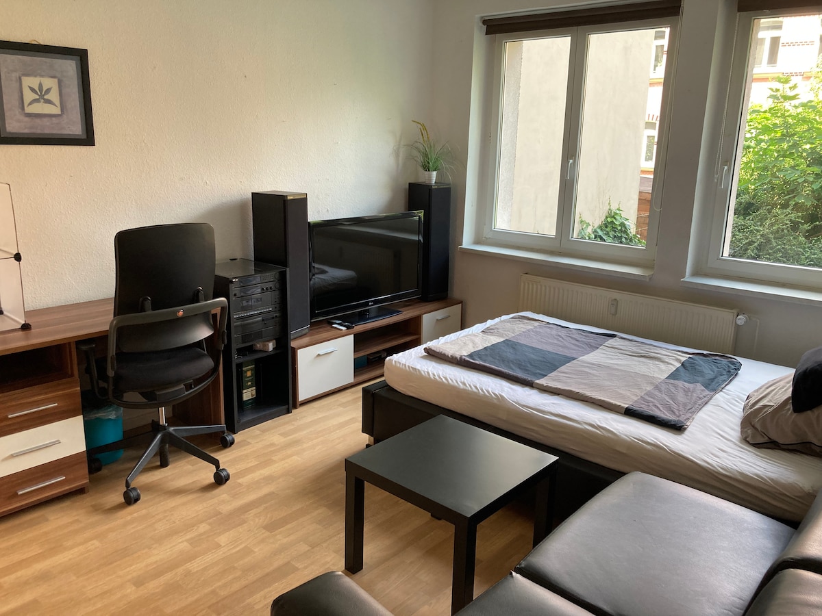 A well-furnished room features a spacious bed with an unmade cover, a two-seater couch, and a small coffee table. A desk with a comfortable chair and a large television are visible. Natural light enters through two windows overlooking a quiet courtyard.