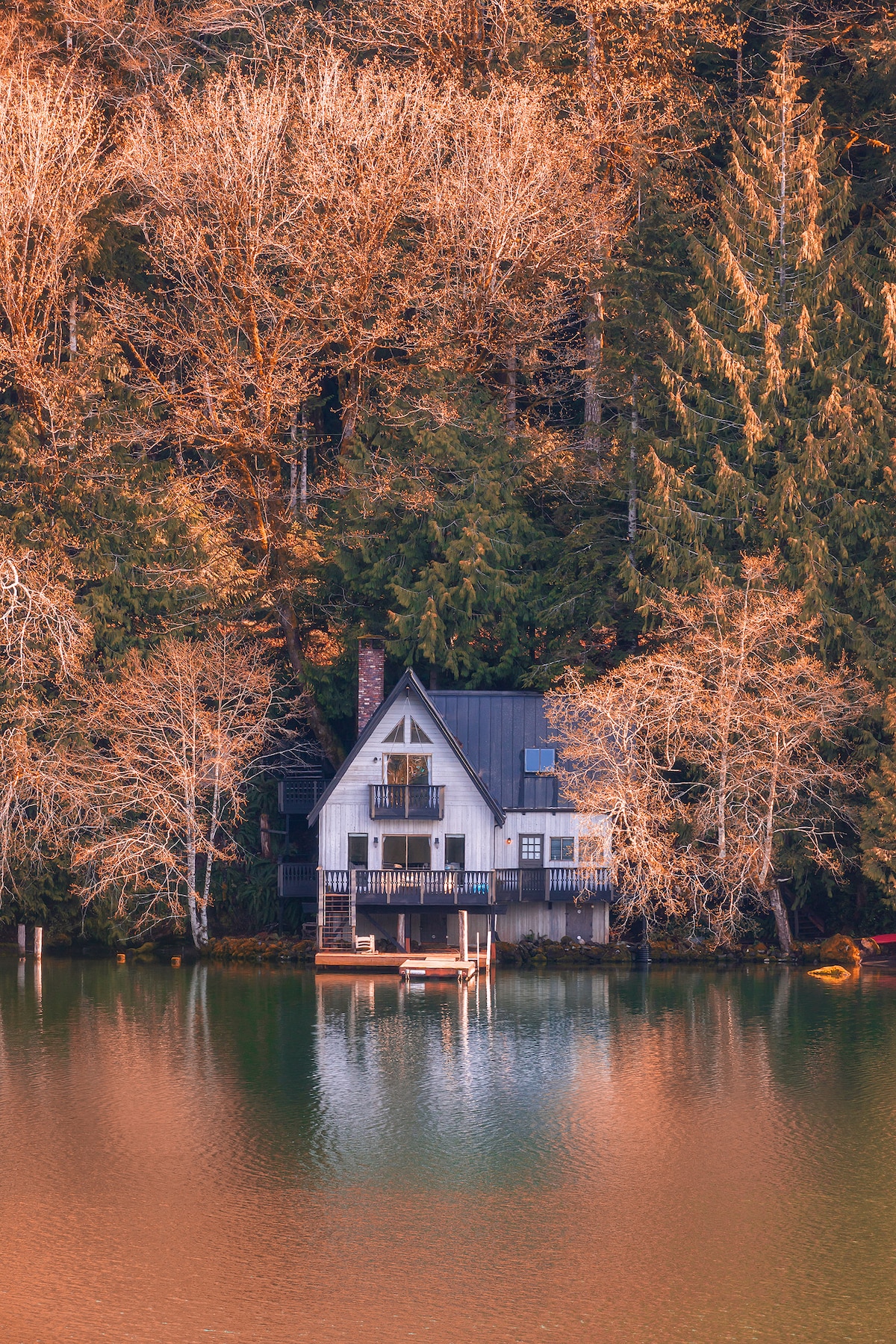 A charming lakefront home, with distinctive architectural features, is nestled among tree-covered slopes. Its reflection on the calm water creates a serene scene, highlighted by the warm tones of the surrounding foliage. The structure is positioned on a private dock, inviting relaxation by the lake.