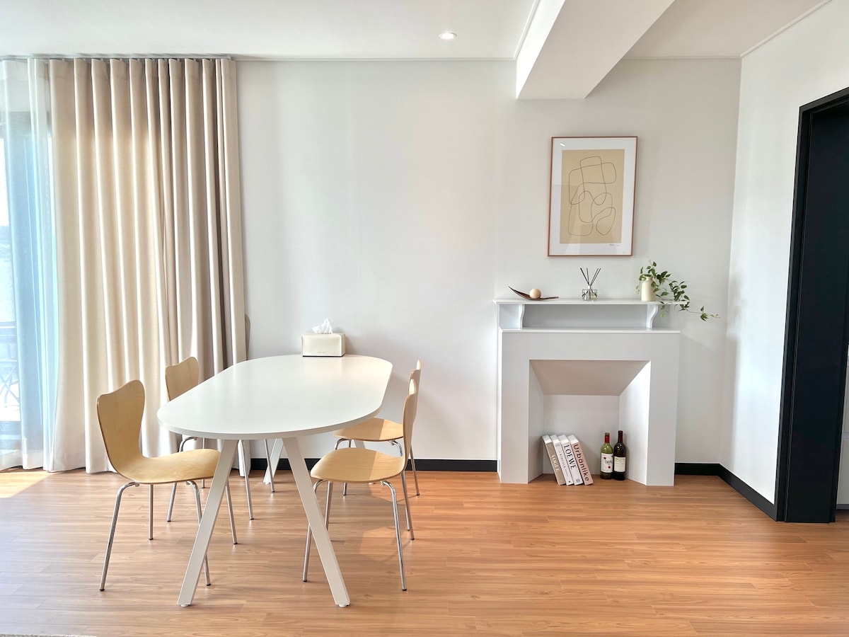 A dining area is presented with a white oval table surrounded by four wooden chairs. A small decorative shelf is visible on the wall, featuring minimalistic decor. Natural light filters through sheer curtains, enhancing the bright and open atmosphere of the space.